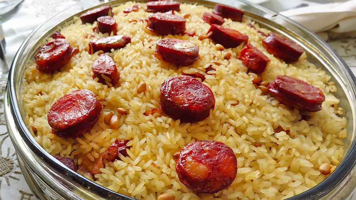 A rustic clay dish filled with golden-brown duck rice, showing tender pieces of duck meat and sliced chouriço mixed throughout the creamy, well-seasoned rice