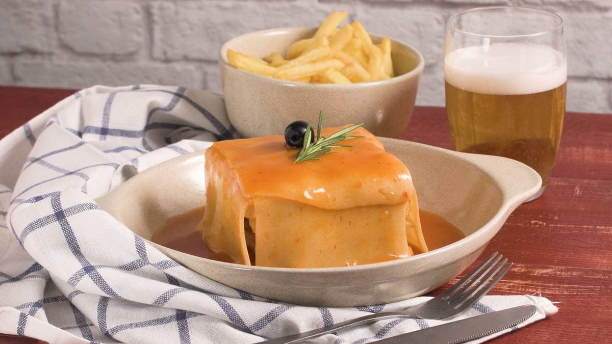 A towering Francesinha sandwich covered in melted cheese and topped with a fried egg, swimming in rich tomato-beer sauce on a white plate