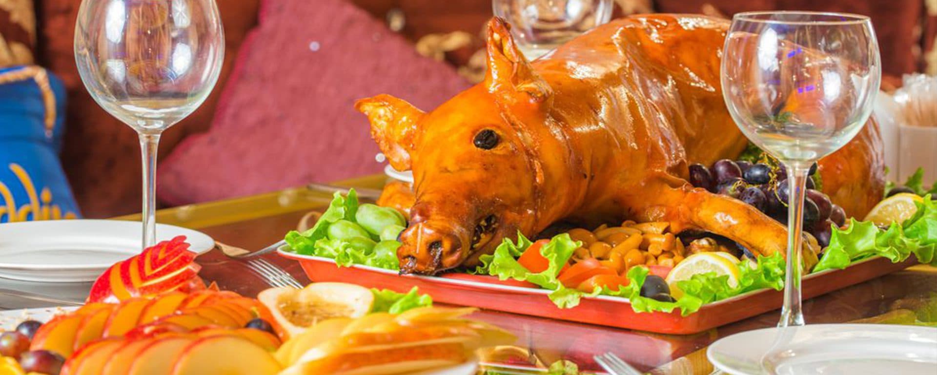 Golden-brown roasted suckling pig with crispy, crackling skin on a large serving platter, garnished with bay leaves and surrounded by roasted potatoes