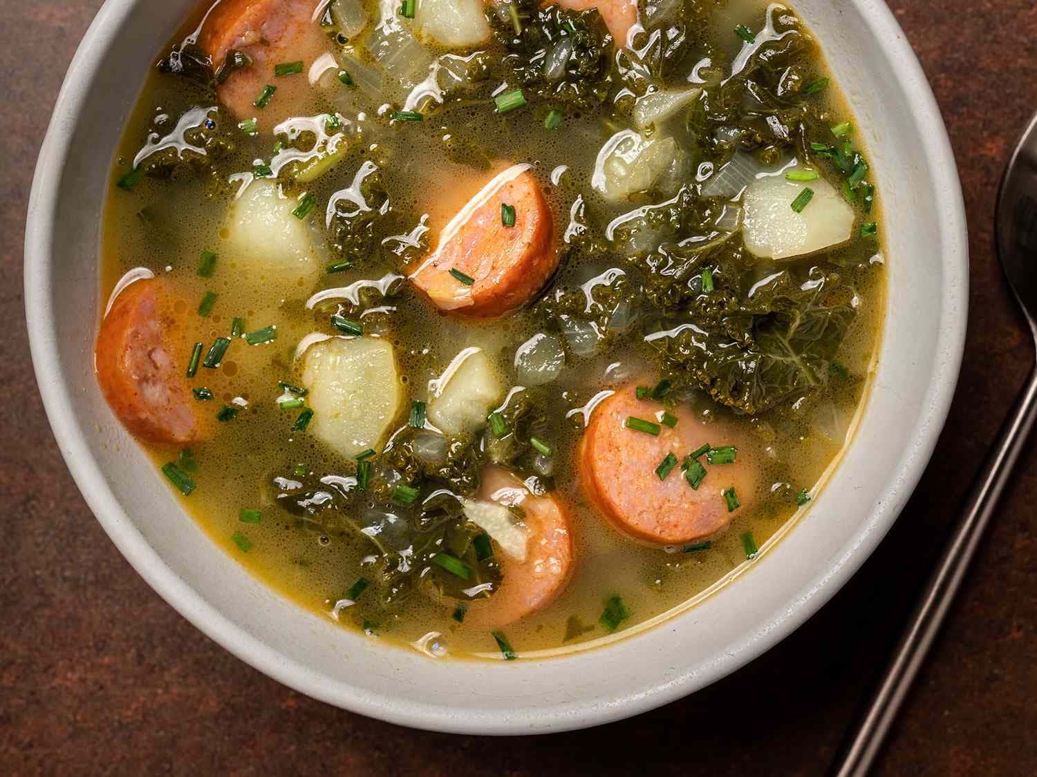 A bowl of vibrant green Caldo Verde soup with thin ribbons of dark green kale, chunks of tender potato, and slices of reddish chouriço sausage, drizzled with golden olive oil
