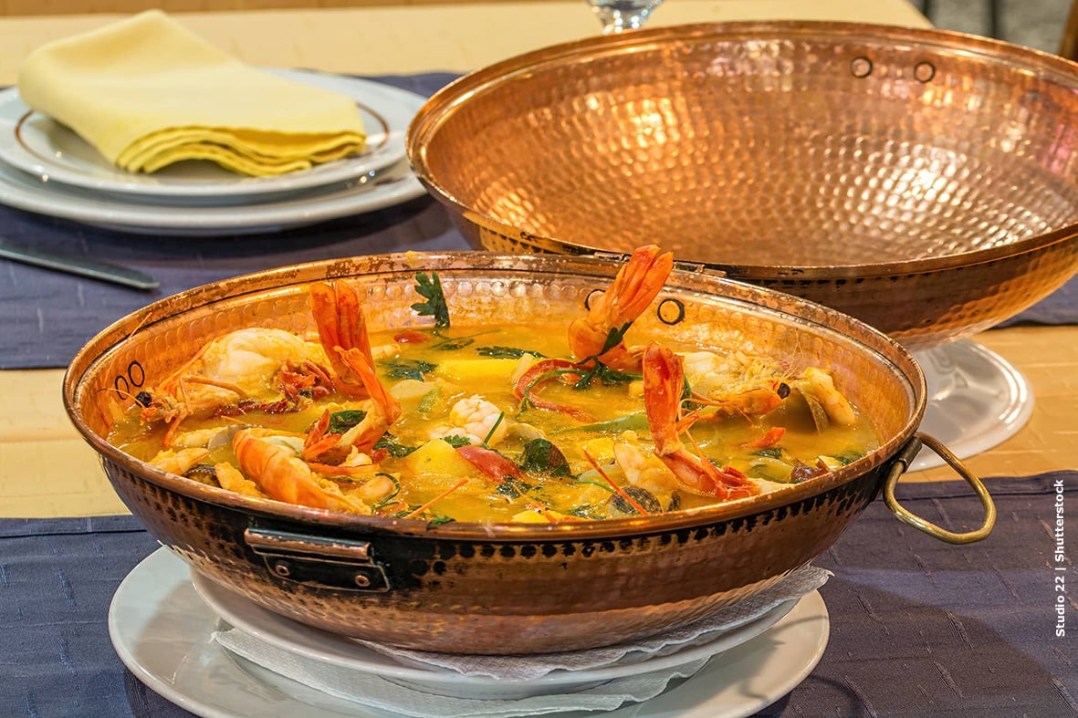 A copper cataplana pot filled with colorful seafood stew featuring prawns, clams, mussels, and monkfish in a rich tomato and pepper sauce, garnished with fresh coriander