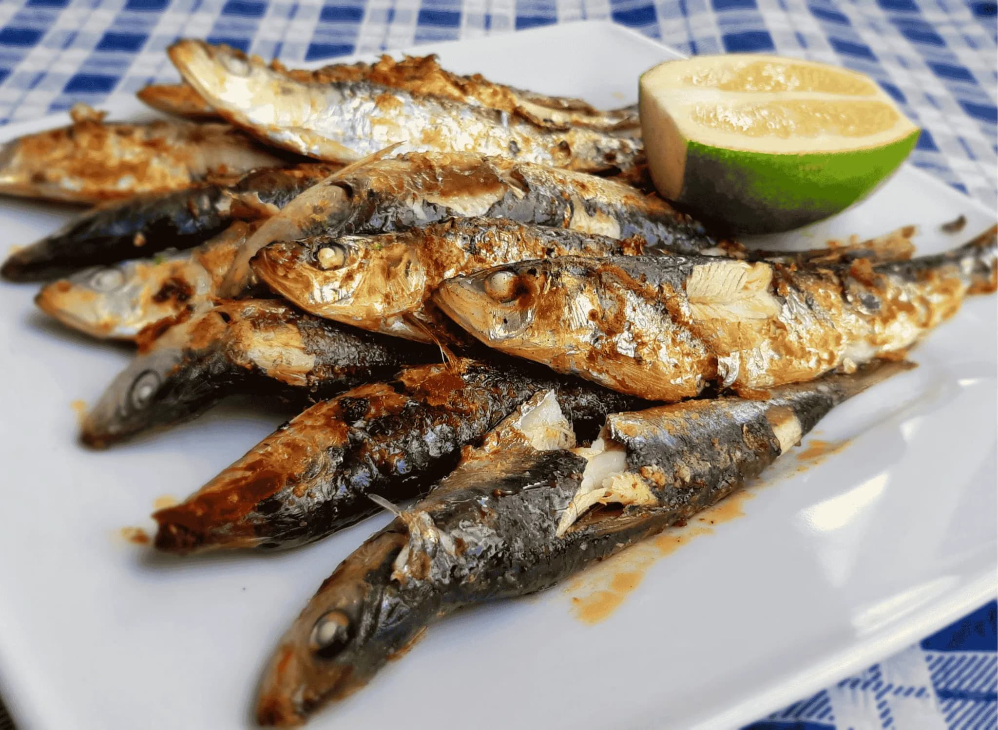 Golden-brown grilled sardines with crispy charred skin arranged on a white platter, garnished with fresh parsley and lemon wedges, with crusty bread in the background