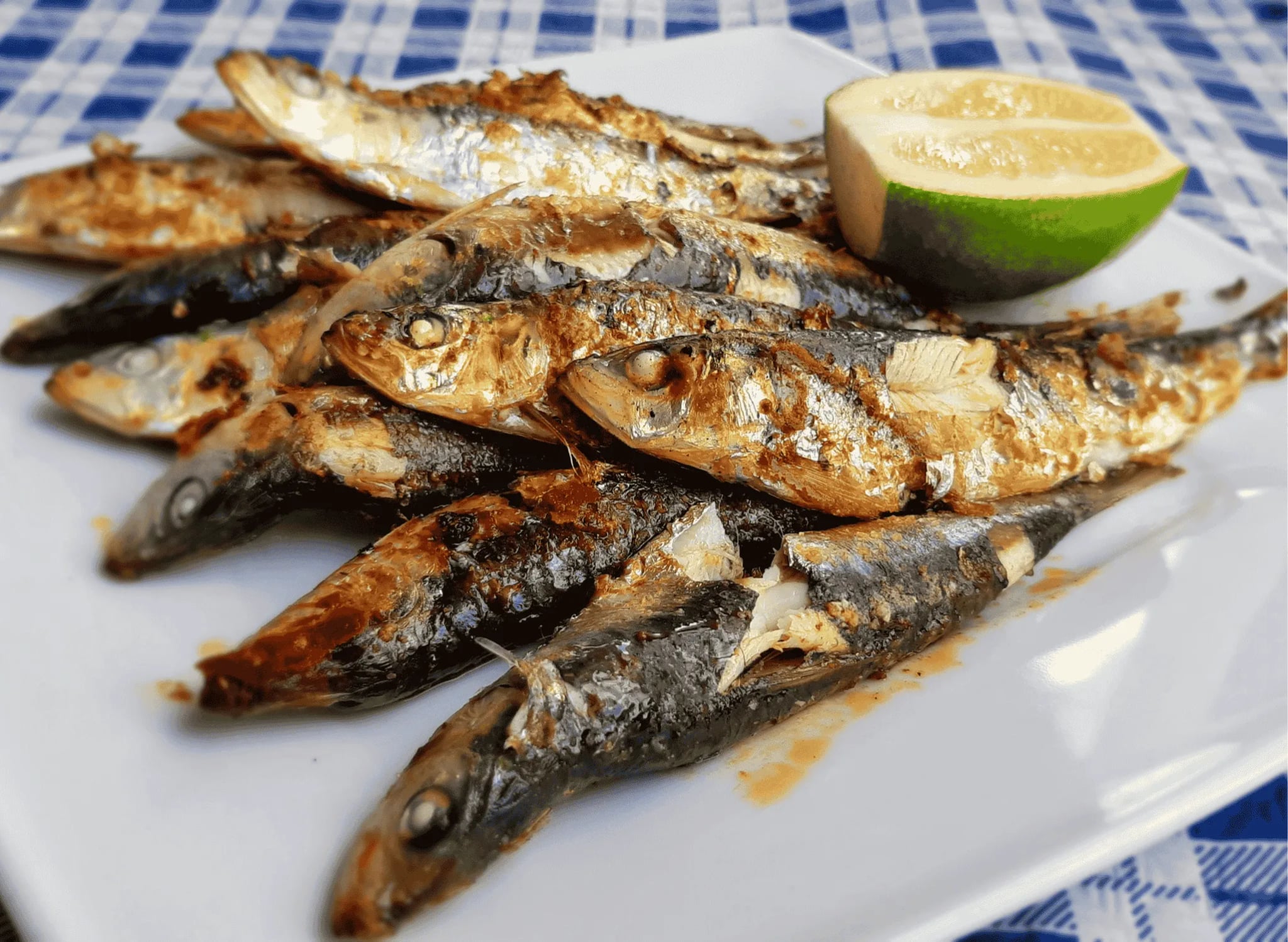 Golden-brown grilled sardines with crispy charred skin arranged on a white platter, garnished with fresh parsley and lemon wedges, with crusty bread in the background
