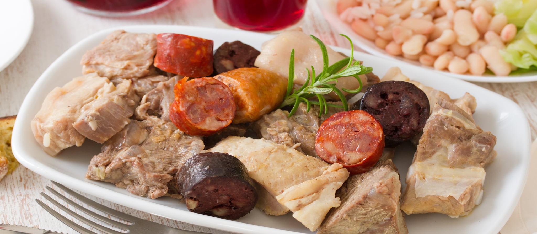 A large platter of Cozido à Portuguesa showing an abundant arrangement of sliced meats, sausages, and colorful vegetables including cabbage, carrots, potatoes, and turnips, with bowls of rich broth alongside
