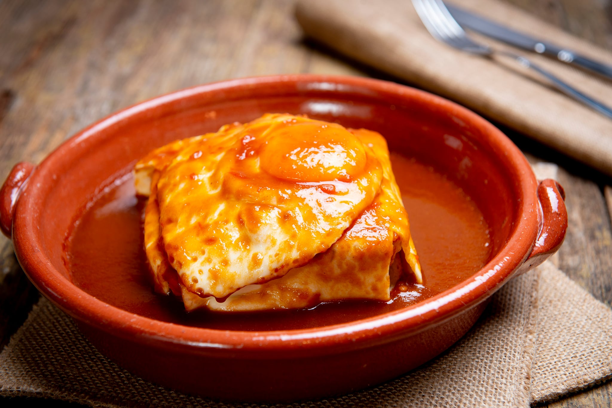 A golden-brown francesinha sandwich covered in melted cheese and rich sauce, served on a white plate with french fries, steam rising from the hot dish